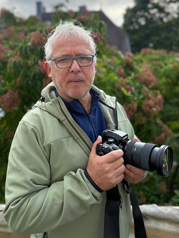 Joachim Schäfer Mann mit Brille und Pflanzen im Hintergrund hält Kamera