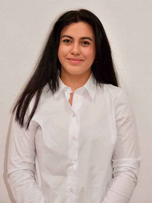 Zehra Açıkgöz Frau mit langen schwarzen Haaren und weißer Bluse vor neutralem Hintergrund
